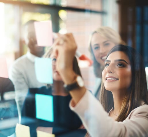 A view of a woman writing on a sticky note on a clear glass window, while her team smiles behind her.